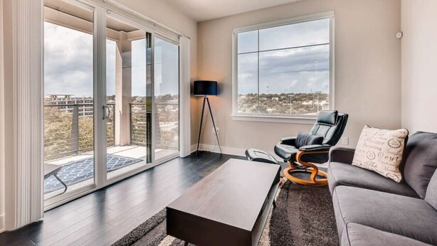 Un salón amplio con un gran ventanal corredero que da a un espacio exterior, una lámpara de pie negra, un sillón de cuero negro, un sofá gris y una mesa de centro de madera.