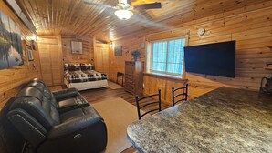 Interior acogedor de una cabaña de troncos con cama, zona de estar con sofá de cuero, mesa de comedor y televisor montado en la pared.