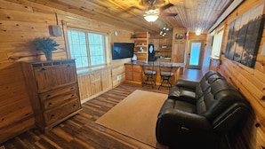 Interior acogedor de una cabaña con piso de madera, un sofá de cuero negro, una mesa de comedor con sillas, una zona de cocina y un ventanal con persianas.
