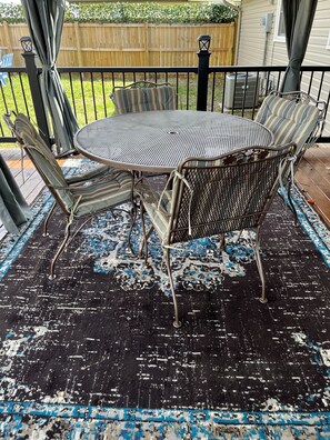 A round metal table with chairs, a black and blue patterned rug, and a wooden fence in the background. - The Key to your Heart (Nixa)