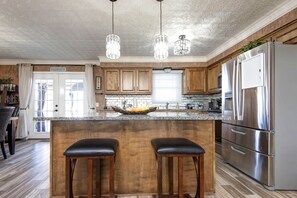 A kitchen with wooden cabinets, a stainless steel refrigerator, a marble countertop, and two bar stools. - The Key to your Heart (Nixa)