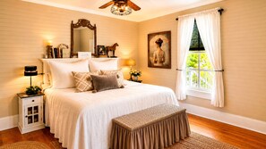 A bedroom with a large bed, a nightstand, a bench, a ceiling fan, a window with curtains, and a painting on the wall.