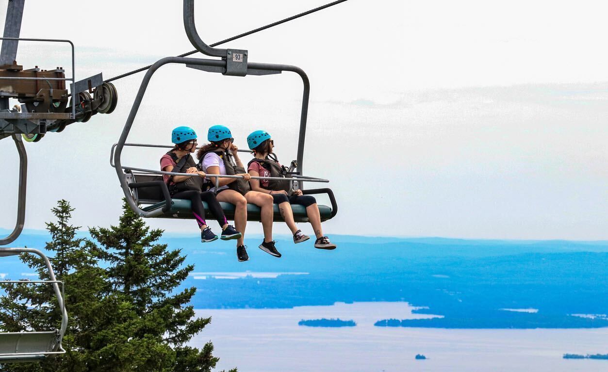 Un télésiège transportant trois passagers portant des casques, offrant une vue panoramique sur un paysage côtier avec des arbres et de l’eau.