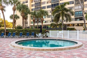 A swimming pool with lounge chairs and palm trees, surrounded by a building with multiple floors and balconies.