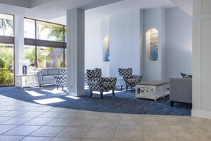 A spacious lobby with a grey sofa, patterned armchairs, and a coffee table. Large windows allow natural light and offer views of palm trees.