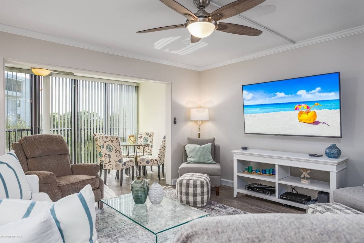 Ein Wohnzimmer mit Deckenventilator, einem Glastisch, einer Couch, einem an der Wand montierten Fernseher und Blick auf den Strand durch das Fenster.