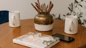 A round wooden table with two white mugs, a smartphone, and a book titled "The Year of the Dog" by Adrianna Adarme.