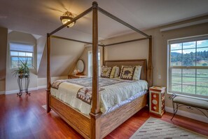 A four-poster canopy bed with a wooden frame, a quilted bedspread, and decorative pillows. There is a small bedside table with a mirror and a plant. The room has wooden flooring and a view of a green landscape through a large window. - Scenic Sanctuary: Monthly Stay, Spectacular Mtn View, Gas Firepit, 10 Min to Downtown Pagosa Springs (Pagosa Springs)