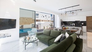 A modern living room with a green sofa, a coffee table, and a TV. There is a dining area with a table and chairs, and a kitchen with a black countertop. A pool is visible through a glass door.