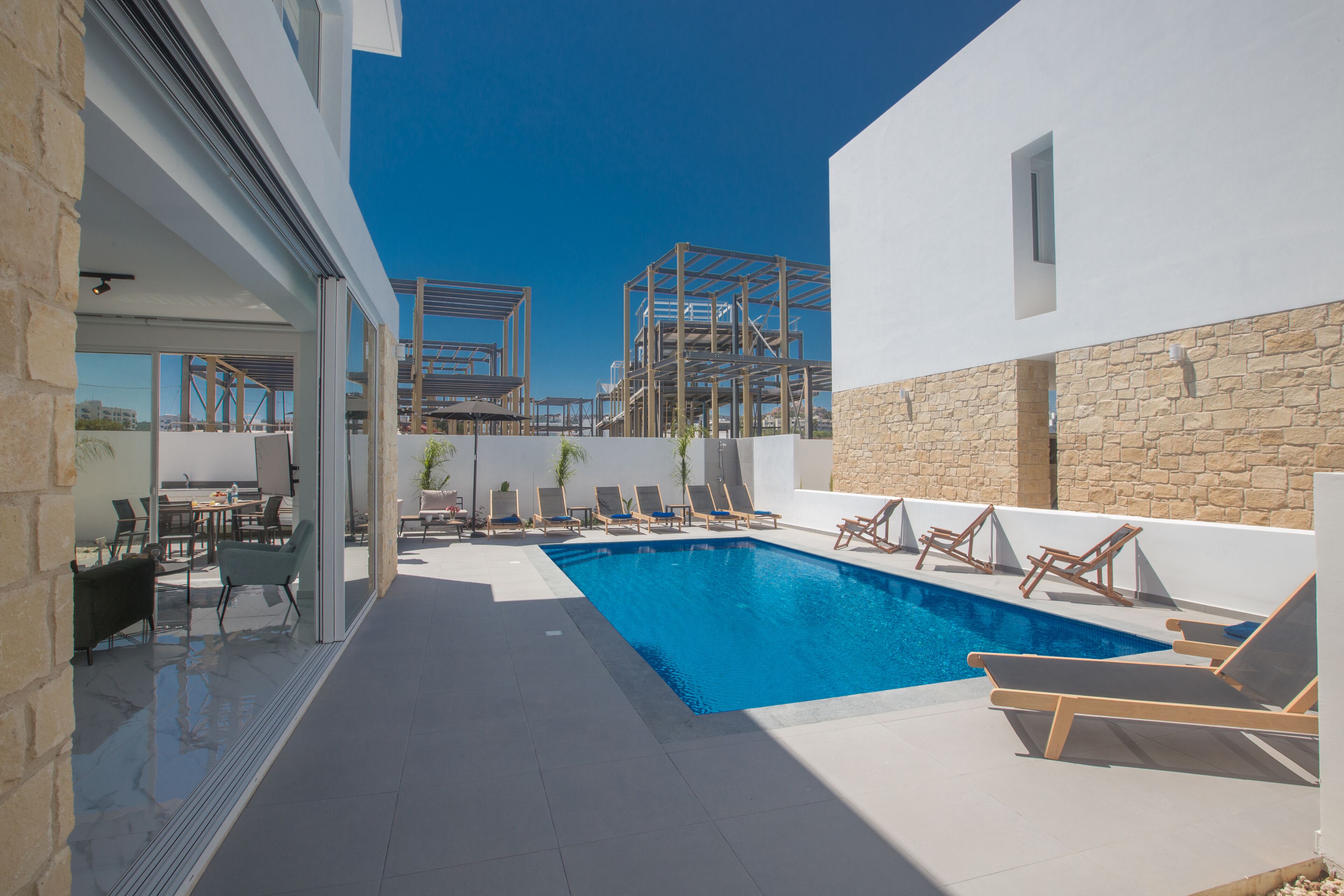 Un área de piscina exterior moderna con sillas de descanso, una pared de piedra y un cielo azul despejado.
