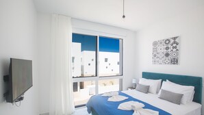 A hotel room with a bed, a TV, a window with curtains, and a view of buildings outside.