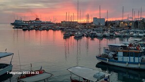 A marina with various boats docked, a large ship in the background, and a sunset in the distance. - Cretan Dawn Apart, Garden, BBQ & near the beach-4 (Heraklion)