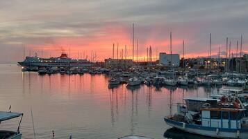 A marina with various boats docked, a large ship in the background, and a sunset in the distance.