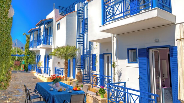 A row of white buildings with blue balconies and doors, a table set for dining, and a clear blue sky. - Cretan Dawn Apart, Garden, BBQ & near the beach-4 (Heraklion)