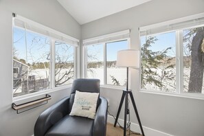 A room with a black leather chair, a standing lamp, and large windows offering a view of trees and a snowy landscape.