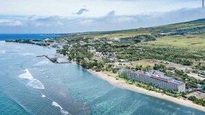 Vista aérea de una zona costera con un hotel, aguas azul cristalino y un paisaje montañoso.