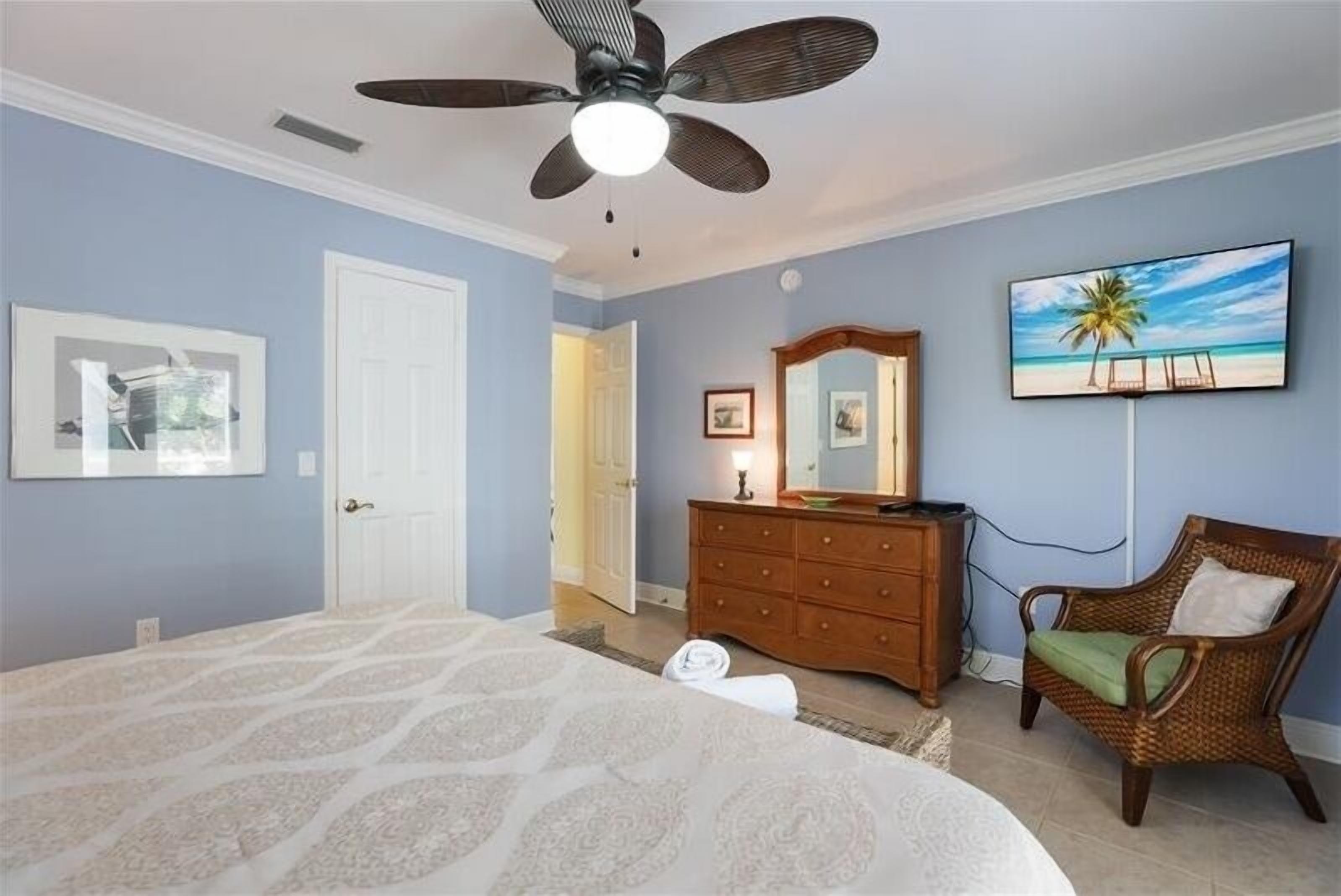 A bedroom with a bed, a dresser, a chair, a ceiling fan, a TV, and a door leading to another room.