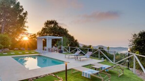 Piscina com espreguiçadeiras e um gazebo, cercada por um gramado bem cuidado e árvores.