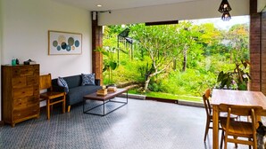 A living room with a sofa, coffee table, wooden cabinet, and a dining table with chairs. There is a large window offering a view of a lush garden. - Ancala Uluwatu (Pecatu)