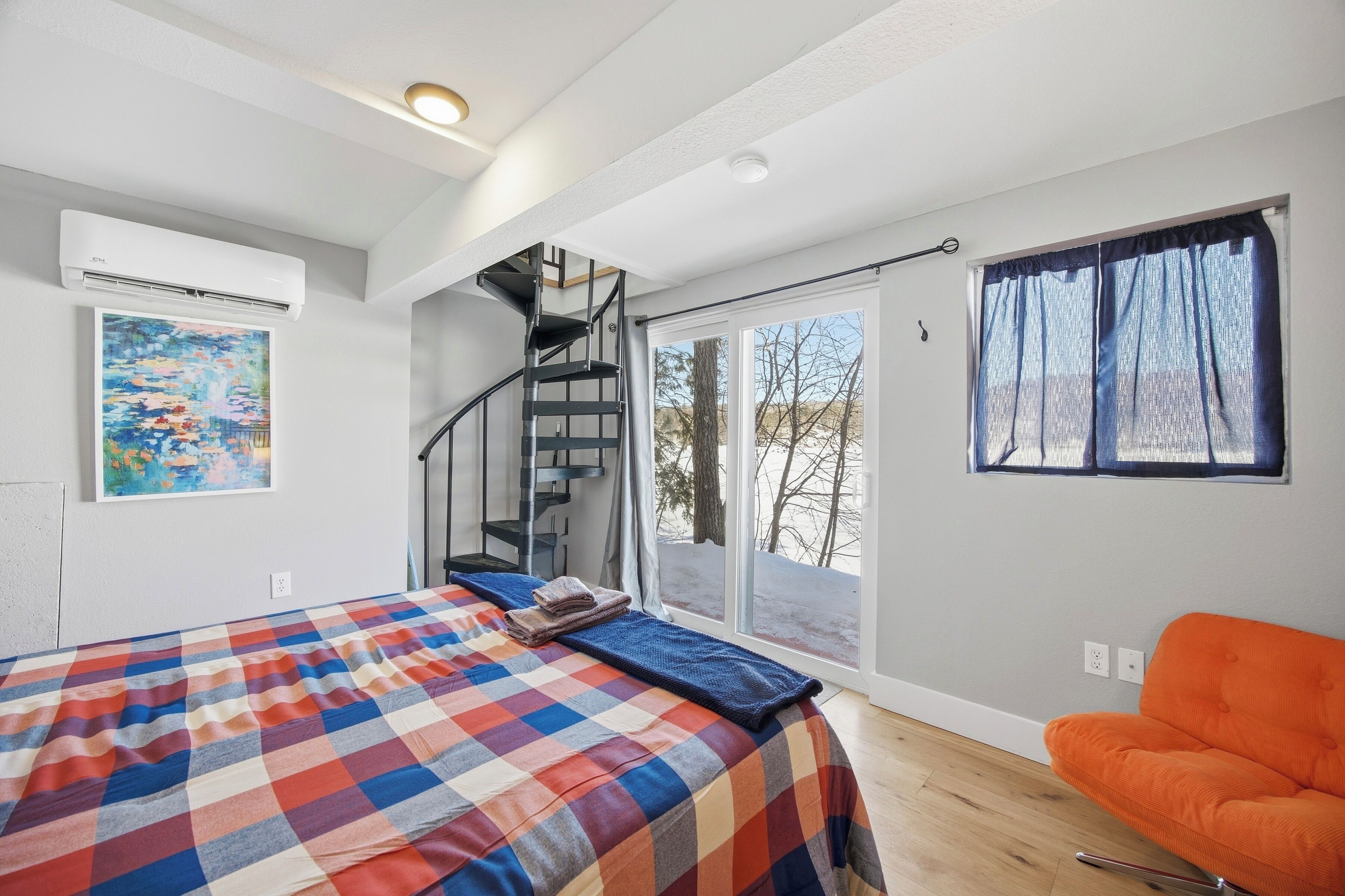 A bedroom with a bed, a chair, a staircase, and a window with a view.