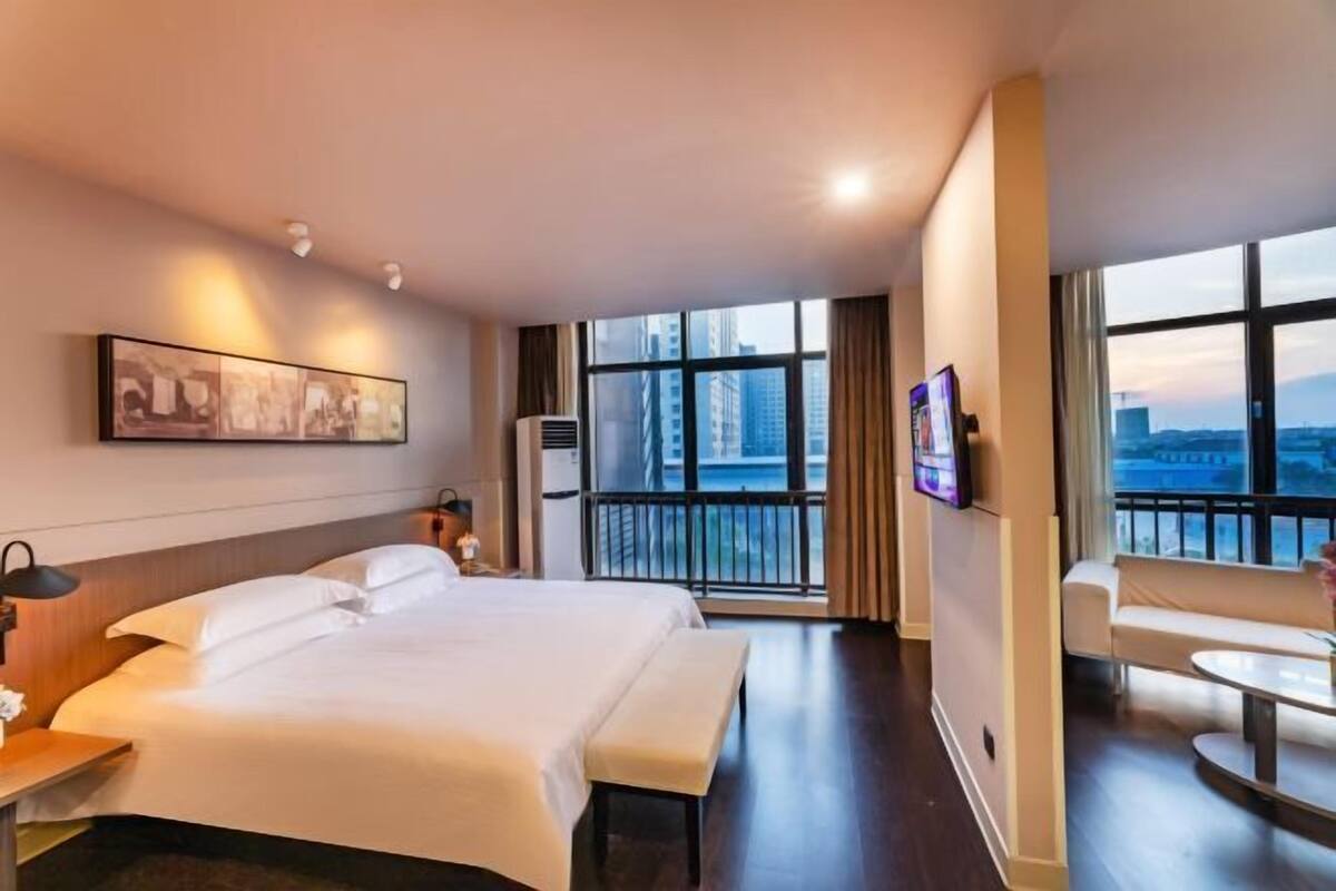Habitación de hotel con una cama grande, un escritorio, una silla y un balcón con vista a la ciudad.