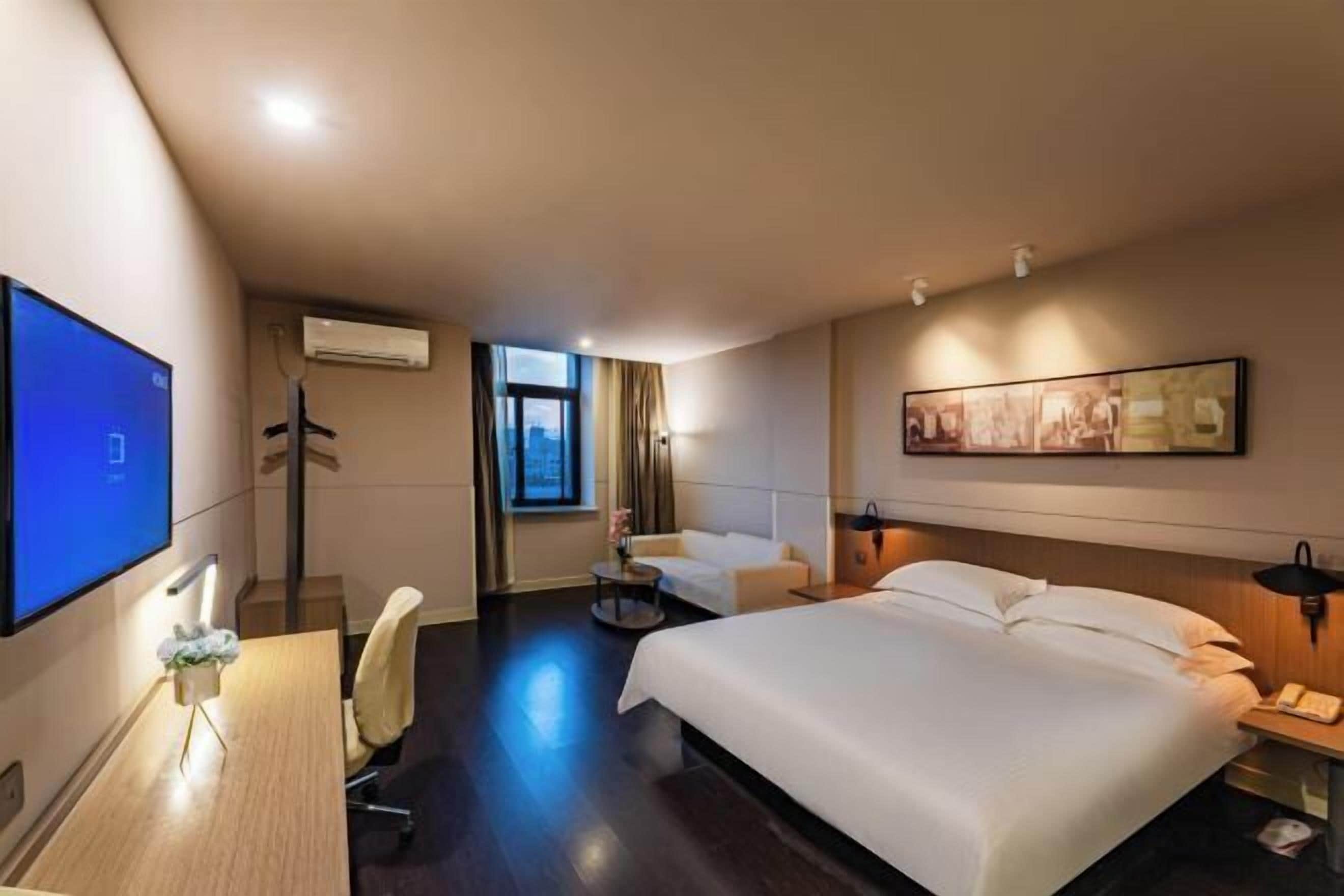 A hotel room with a large bed, a desk with a computer, a chair, a small round table with a vase, and a window with curtains.