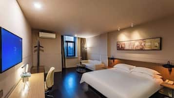 A hotel room with a large bed, a desk with a computer, a chair, a small round table with a vase, and a window with curtains.