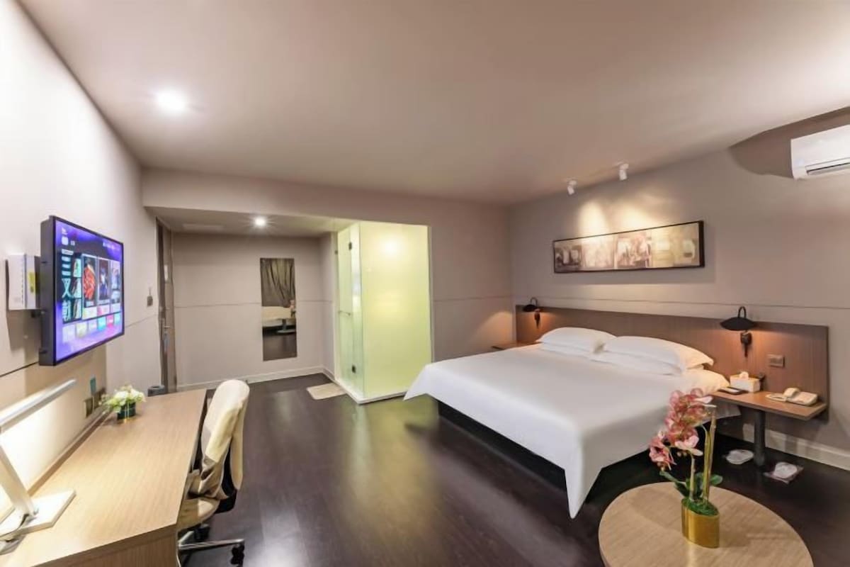 Habitación de hotel con cama, mesita de noche, lámpara, televisor, escritorio, silla y una pequeña mesa con flores.