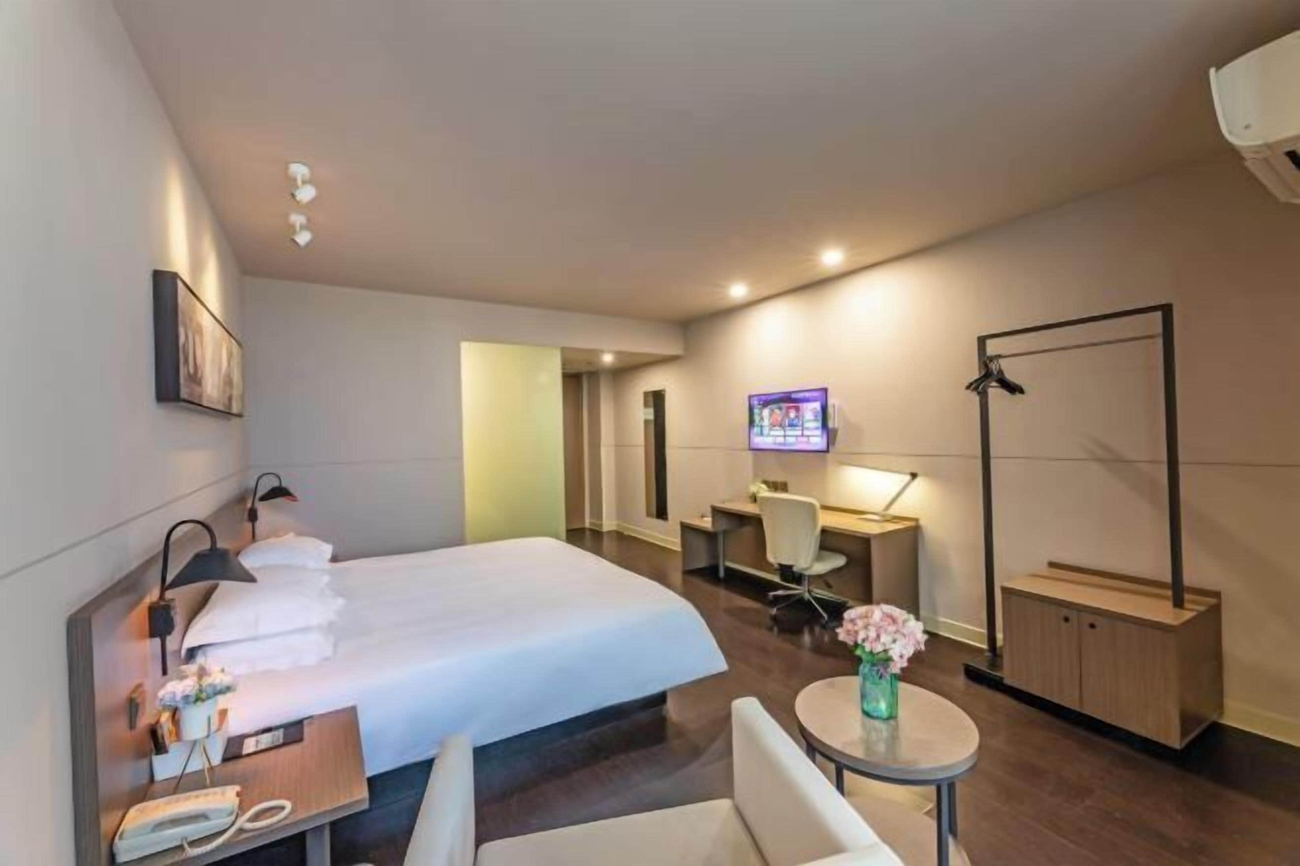 A hotel room with a bed, bedside table, TV, desk, chair, and a small table with flowers.