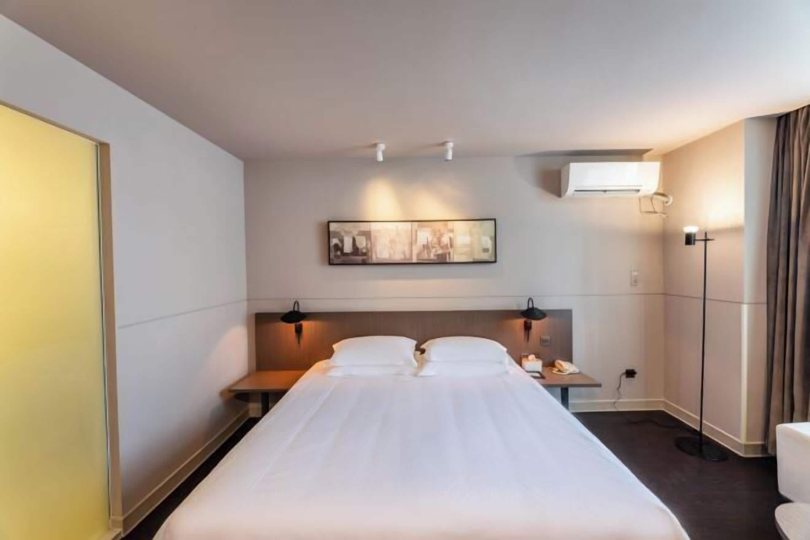 Quarto de hotel com cama, mesas de cabeceira, ar condicionado fixado na parede, abajur e um quadro emoldurado.