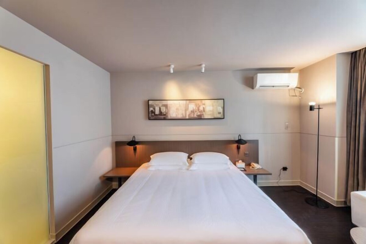 Una habitación de hotel con una cama, mesitas de noche, aire acondicionado en la pared, una lámpara de pie y un cuadro enmarcado.