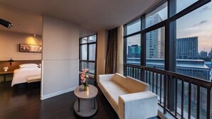 A hotel room with a bed, a sofa, a small table with flowers, and large windows offering a city view.