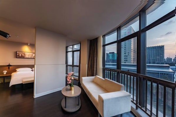 Habitación de hotel con cama, sofá, una pequeña mesa con flores y ventanas amplias con vistas a la ciudad.