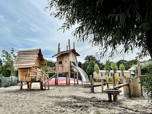 Une structure de jeu en bois, dotĂ©e dâun toboggan et dâĂ©lĂ©ments dâescalade, entourĂ©e de sable et de verdure.