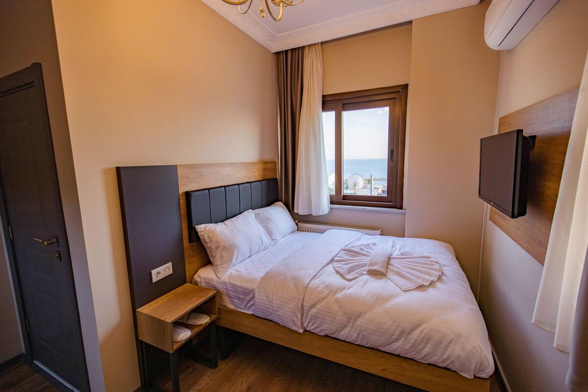 A neatly made bed with a wooden headboard, a bedside table, a TV mounted on the wall, and a window with curtains.