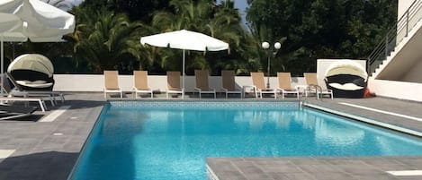 A swimming pool with lounge chairs and umbrellas, surrounded by palm trees and a modern building.