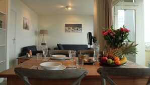 A dining area with a table set for two, a sofa in the background, and a fruit bowl on the table. - Cosy apartment in Noordwijk aan Zee with balcony (Noordwijk aan Zee)