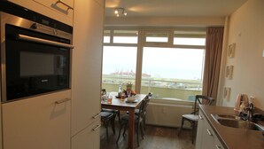 A kitchen with a Siemens oven, a dining table with chairs, a view of the sea through large windows, and a radiator under the table. - Cosy apartment in Noordwijk aan Zee with balcony (Noordwijk aan Zee)