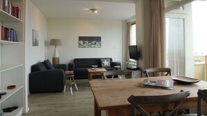 A modern living room with a sofa, coffee table, and dining table. There is a bookshelf, a lamp, and a painting on the wall. - Cosy apartment in Noordwijk aan Zee with balcony (Noordwijk aan Zee)