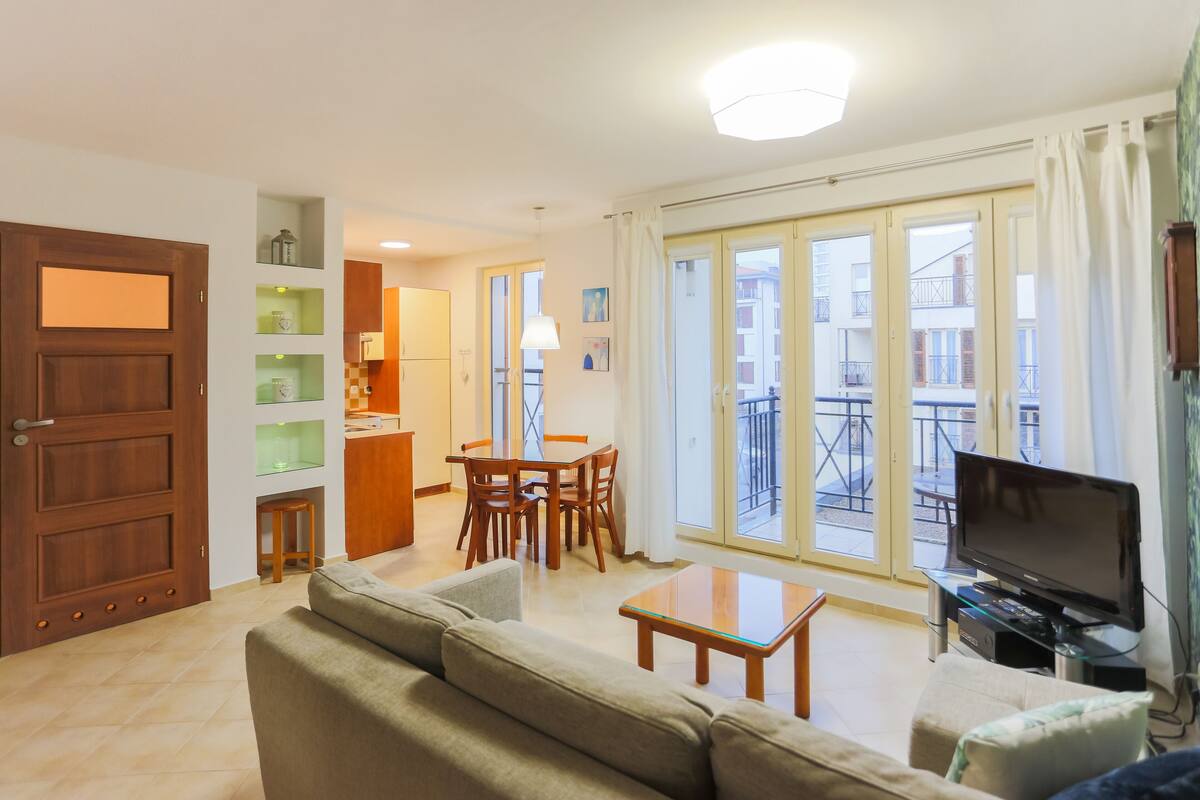 A living room with a sofa, a dining table, and a TV. There is a balcony with a view of buildings.