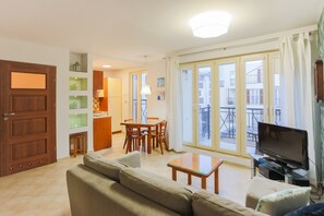 A living room with a sofa, a dining table, and a TV. There is a balcony with a view of buildings.