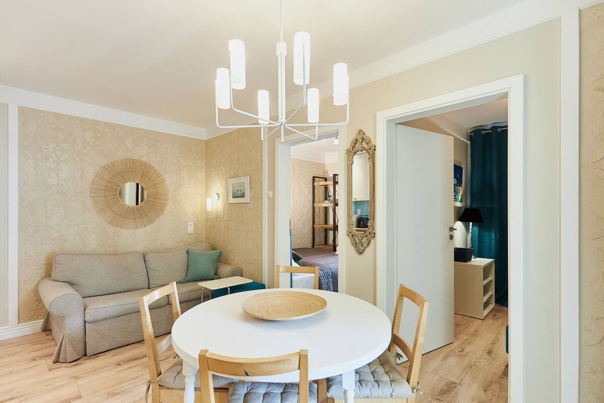 A dining area with a round table, chairs, a chandelier, a sofa, and a mirror on the wall.