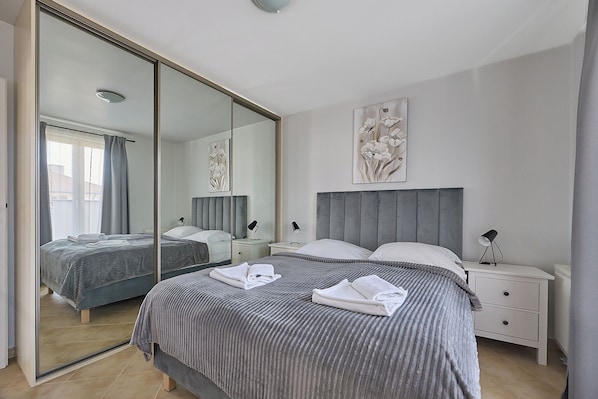 A bedroom with a large mirror, a bed with two pillows, a bedside table with a lamp, and a wall-mounted artwork.