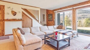 A spacious living room with a large sectional sofa, a glass coffee table, and a view of a mountainous landscape through large sliding glass doors. - Luxury Hunter Mountain View Townhome -Pool-Fire pit-Walk to Scribner’s-Sleeps 10 (Hunter)