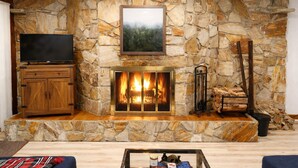 A stone fireplace with a TV above it, a wooden cabinet to the left, and a coffee table in the foreground. - Luxury Hunter Mountain View Townhome -Pool-Fire pit-Walk to Scribner’s-Sleeps 10 (Hunter)