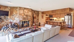 A modern living room with a stone fireplace, a large sofa, and a kitchen area with wooden cabinets and stainless steel appliances. - Luxury Hunter Mountain View Townhome -Pool-Fire pit-Walk to Scribner’s-Sleeps 10 (Hunter)