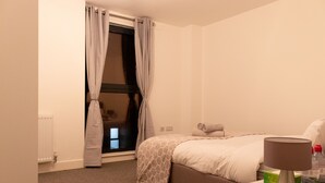 Ein Schlafzimmer mit Bett, Nachttisch, Lampe und einem Fenster mit Vorhängen.