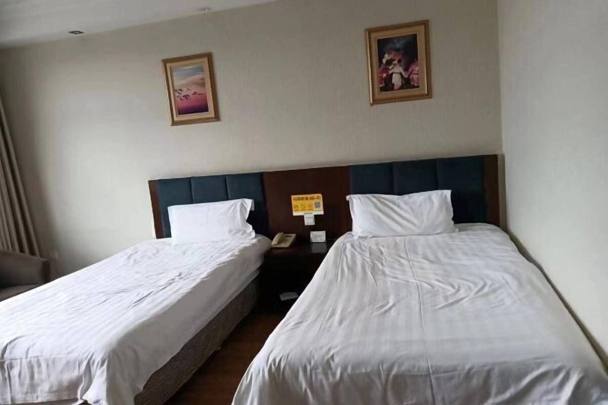 A hotel room with two single beds, a wooden headboard, a nightstand with a phone, and two framed pictures on the wall.