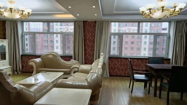 Quarto de hotel com sofá, mesa de jantar e cadeiras. Grandes janelas com cortinas e um candelabro à vista.