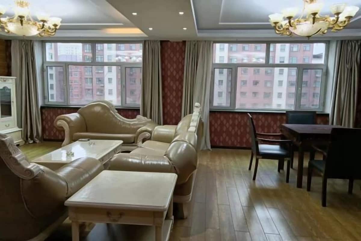 A hotel room with a sofa set, a dining table, and chairs. There are large windows with curtains, and a chandelier is visible.